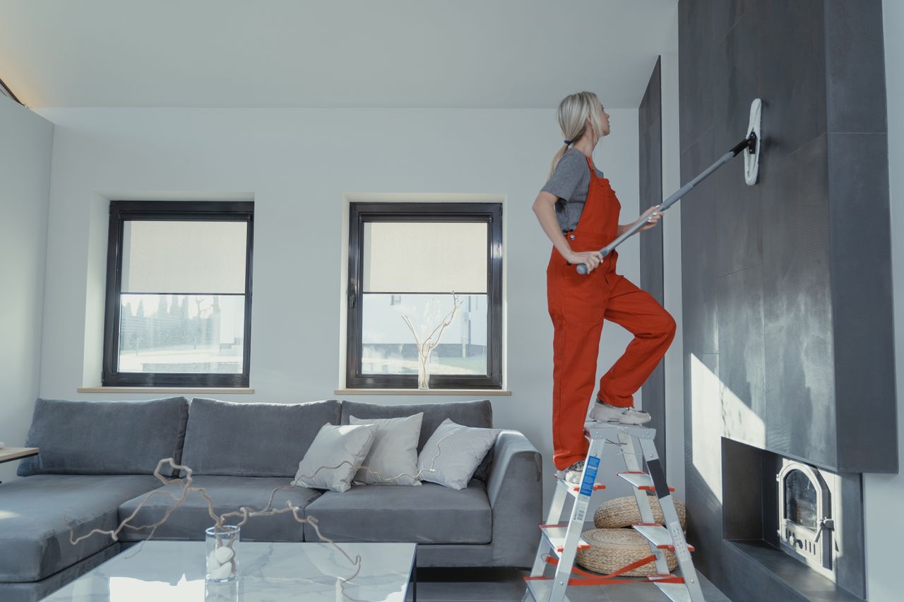 Une femme en salopette nettoie un mur dans un salon élégant, contribuant à l’hygiène de la maison.