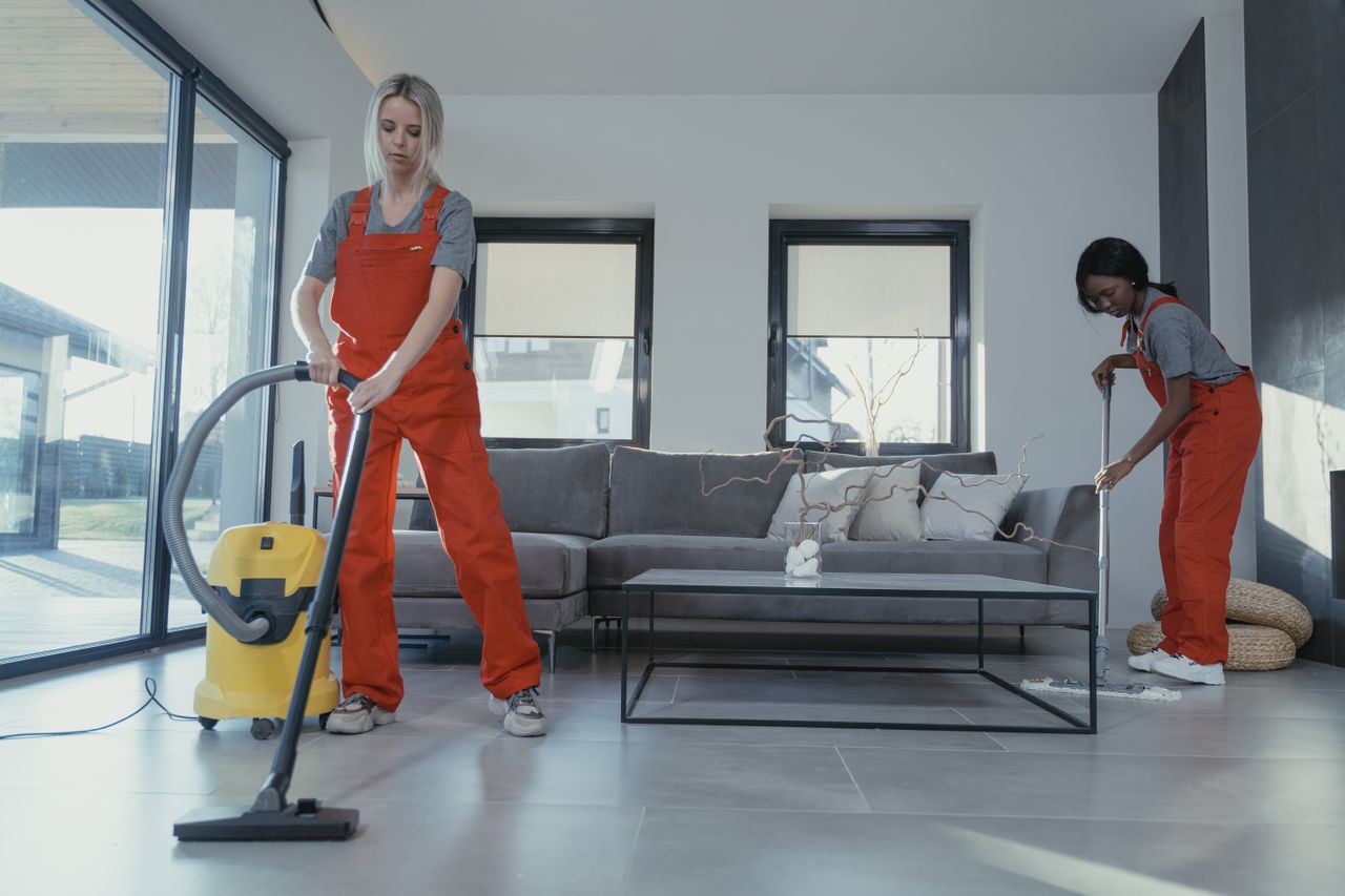 services-01 Two women in red overalls are vacuuming and sweeping a contemporary living room.